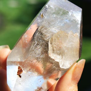May include: A clear quartz crystal with a double terminated point and inclusions of golden rutile. The crystal is approximately 4 inches tall and has a natural, raw appearance.