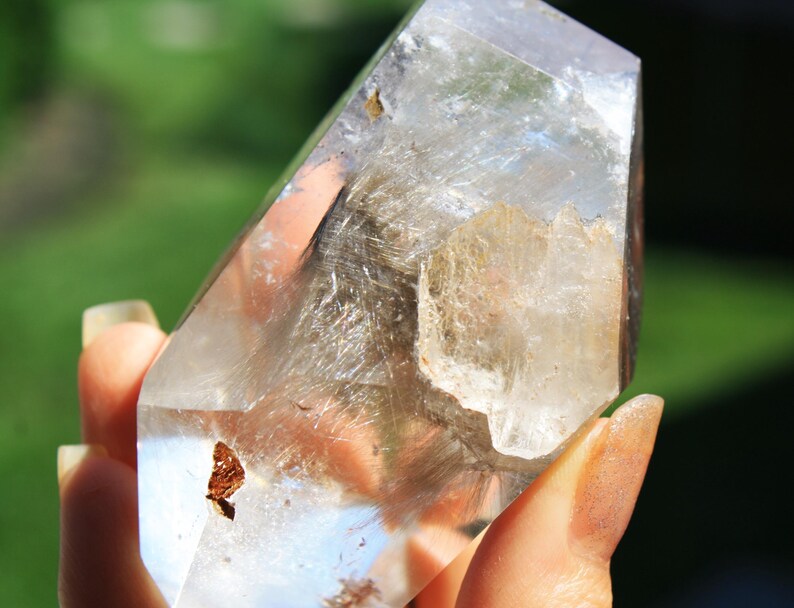 May include: A clear quartz crystal with a double terminated point and inclusions of golden rutile. The crystal is approximately 4 inches tall and has a natural, raw appearance.