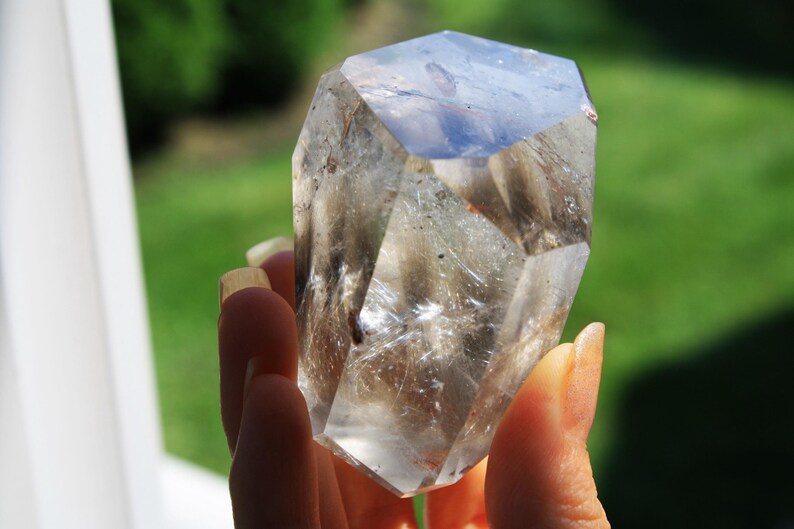 May include: A clear quartz crystal with a smoky gray hue. The crystal is faceted and has a pointed tip. It is being held in a hand.