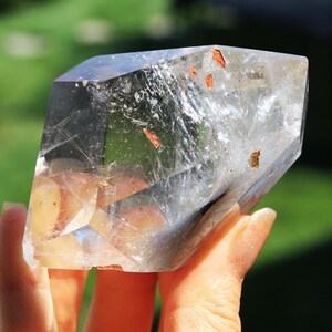 May include: A clear quartz crystal with a double termination and inclusions of red and gold. The crystal is held in a hand.