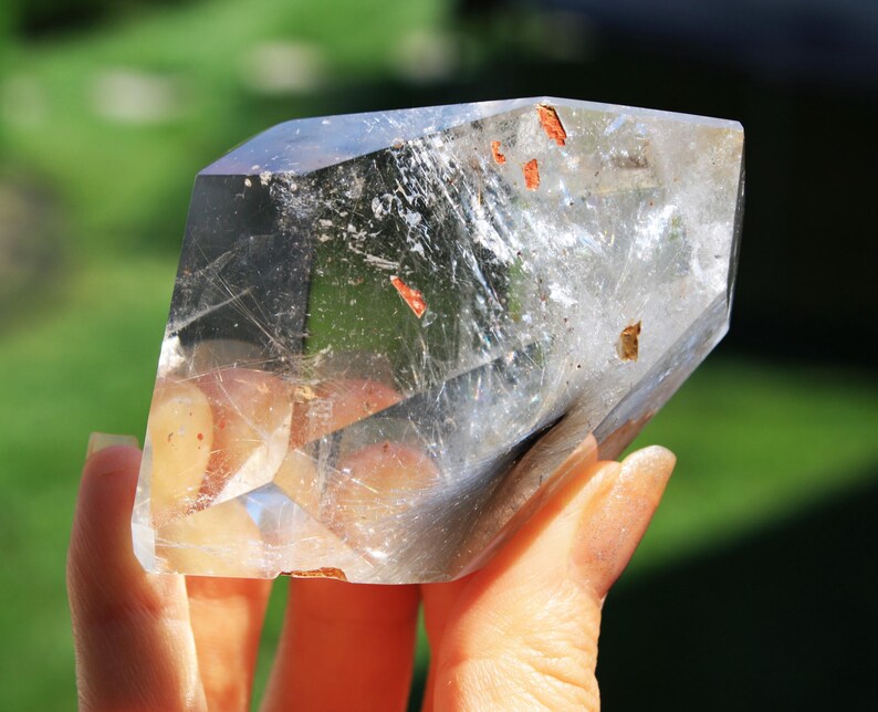 May include: A clear quartz crystal with a double termination and inclusions of red and gold. The crystal is held in a hand.