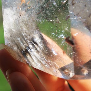 May include: A clear quartz crystal with inclusions of white and black fibers. The crystal is being held in a hand.