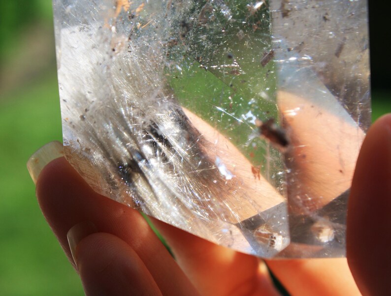 May include: A clear quartz crystal with inclusions of white and black fibers. The crystal is being held in a hand.