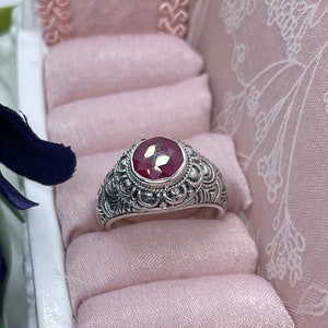 May include: A silver ring with a large, red gemstone set in a floral design. The ring is displayed on a pink velvet ring holder.