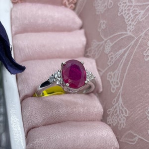 May include: A silver ring with a large oval ruby gemstone surrounded by smaller diamonds. The ring is displayed in a pink velvet ring box.