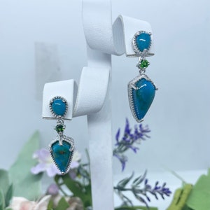 Op de afbeelding: Een paar zilveren oorhangers met turquoise en groene edelstenen. De oorbellen hebben een traanvormige turquoise steen met een zilveren zetting en een kleinere groene edelsteen erboven.