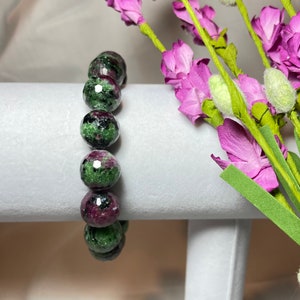 May include: A green and pink gemstone bracelet with faceted beads. The bracelet is on a white display stand.