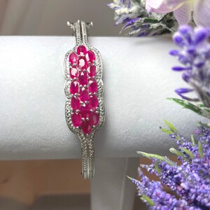 Könnte beinhalten: Ein silbernes Armreif-Armband mit einem Cluster aus ovalen Rubin-Edelsteinen, die in einem dekorativen Design gefasst sind. Das Armband befindet sich auf einer weißen Oberfläche mit lila Blumen.