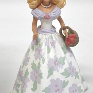 May include: A decorative figurine of a woman in a white floral dress, holding a basket of red fruit. The figurine has blonde hair, blue eyes, and a pearl necklace. She wears a white hat with a pink flower.