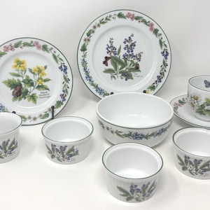 May include: A white porcelain dinnerware set featuring floral and butterfly designs. The set includes plates, bowls, and cups with a green trim. The plates display botanical illustrations of yellow and purple flowers.