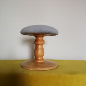 Puede incluir: Un soporte de sombrero de madera con una parte superior acolchada de color gris claro. El soporte tiene una base redonda y un tallo de madera torneada. El soporte está sobre una superficie amarilla.