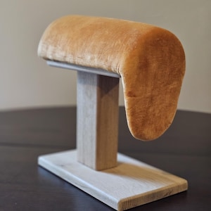 Könnte beinhalten: Ein Schmuckständer mit rechteckiger Holzbasis und vertikaler Holzstütze. Die Oberseite hat eine geschwungene, gepolsterte Auflage, die mit weichem, goldbraunem Samt bedeckt ist. Zum Halten von Armbändern oder Uhren. Ca. 25 cm hoch.
