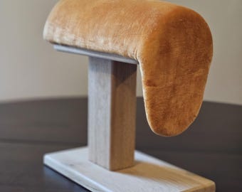 May include: A jewelry display stand featuring a rectangular wooden base and a vertical wooden support. The top has a curved, padded rest covered in a soft, golden-brown velvet material. The stand is designed to hold bracelets or watches.