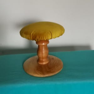 May include: A small, decorative stool with a round, mustard-yellow velvet cushion. The cushion sits atop a turned wooden pedestal and a circular base. The wood has a natural finish, and the stool is set against a teal surface.