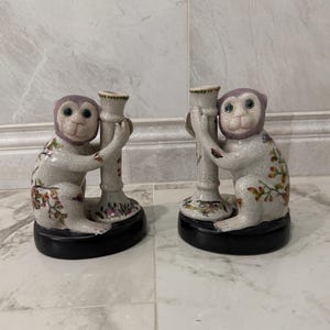 May include: Pair of ceramic monkey candle holders. The monkeys are white with purple faces and floral accents. Each monkey holds a cream-colored candle holder with a black base. Decorative home decor.