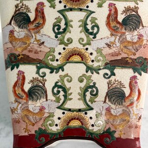 May include: A ceramic vase with a cream-coloured crackle glaze finish, featuring a repeating pattern of roosters and hens in shades of red, orange, and green. The design includes ornate scrollwork and a deep red border at the top and bottom.