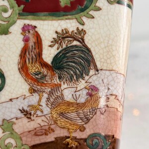 May include: A ceramic vessel with a cream-coloured crackle glaze finish featuring a detailed illustration of a rooster and a hen. The scene includes green scrollwork borders and a deep red top section, showcasing intricate painted feathers.