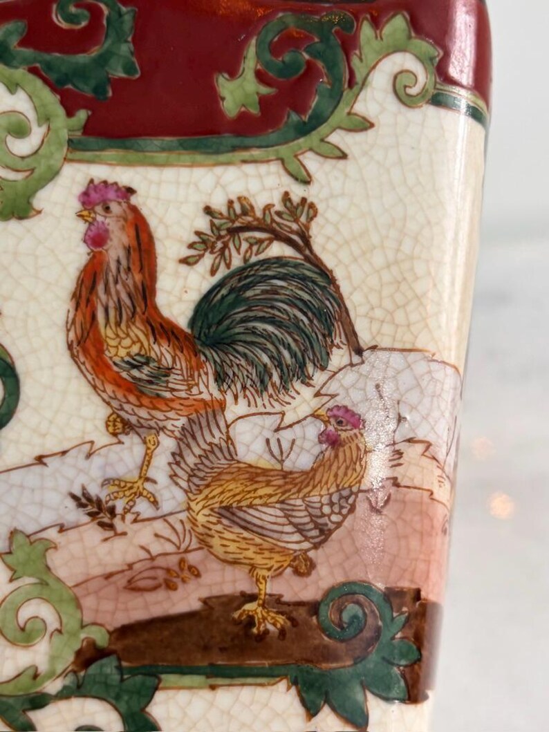 May include: A ceramic vessel with a cream-coloured crackle glaze finish featuring a detailed illustration of a rooster and a hen. The scene includes green scrollwork borders and a deep red top section, showcasing intricate painted feathers.