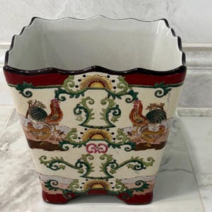 May include: A square ceramic planter with a scalloped rim and a cream-colored crackle glaze finish. The exterior features a repeating pattern of roosters, green scrolling vines, and yellow sunburst motifs, framed by deep red bands at the top and base.