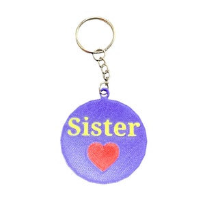 Pode incluir: Chaveiro roxo com um anel e uma corrente prateados. O chaveiro tem um charme redondo com a palavra "Irmã" em letras amarelas e um coração vermelho.