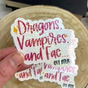 May include: A white sticker with pink glitter and text that reads "Dragons, Vampires, and Fae... Oh my!"