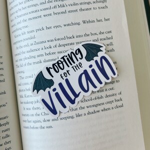 May include: A white sticker with the text "Rooting for the Villain" in a dark blue script. The sticker has a bat wing design in dark teal at the top. The sticker is placed on an open book.