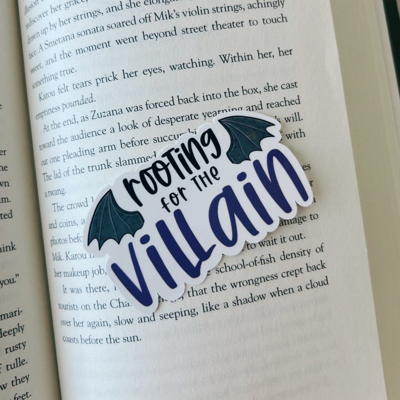 May include: A white sticker with the text "Rooting for the Villain" in a dark blue script. The sticker has a bat wing design in dark teal at the top. The sticker is placed on an open book.