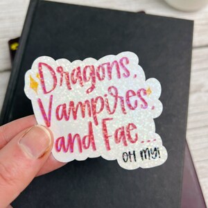 May include: A white sticker with pink glitter lettering that reads "Dragons, Vampires, and Fae... Oh my!"