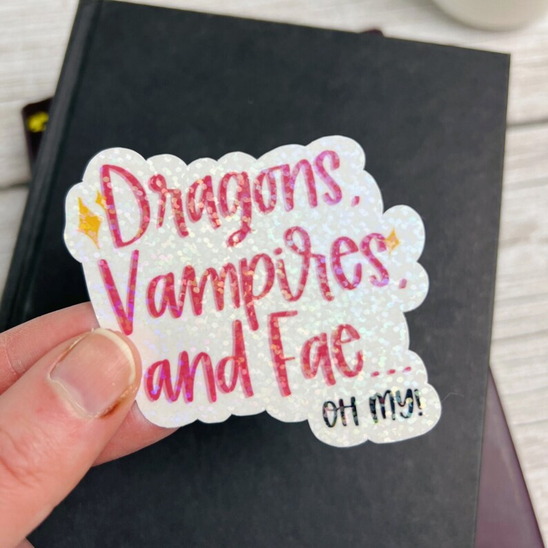 May include: A white sticker with pink glitter lettering that reads "Dragons, Vampires, and Fae... Oh my!"