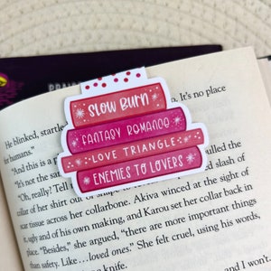 May include: A pink and red bookmark with a stack of books design. The bookmark has the text "Slow Burn", "Fantasy Romance", "Love Triangle", and "Enemies to Lovers" on it.