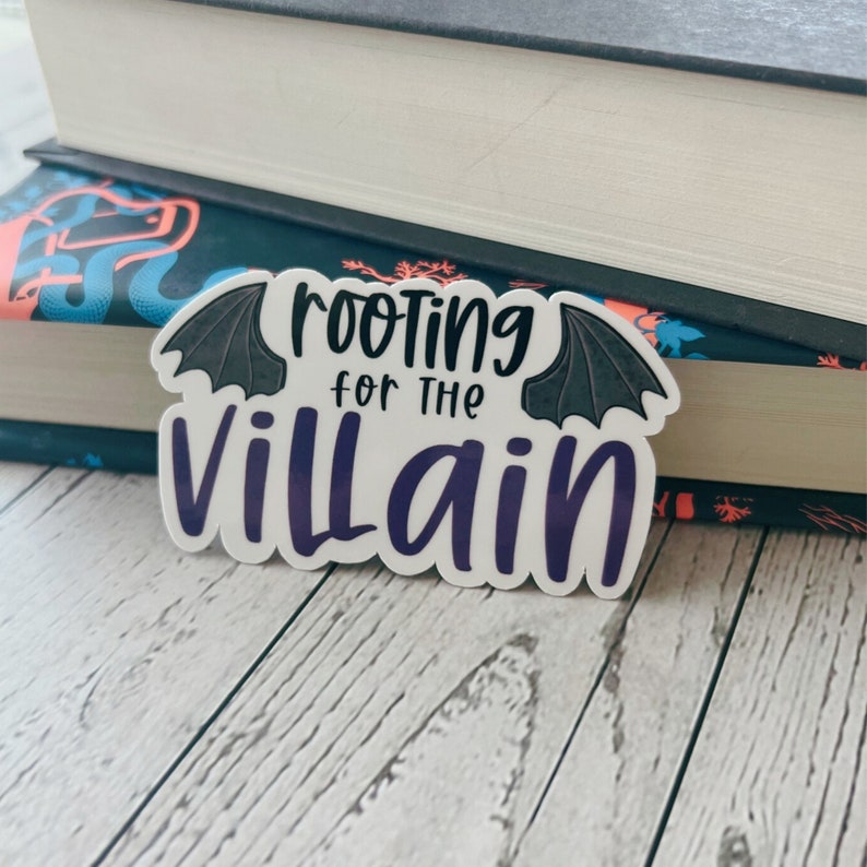 May include: A white sticker with the text "rooting for the villain" in black and purple, with bat wing accents. The sticker is placed on top of two books with dark covers and a light wood grain surface.