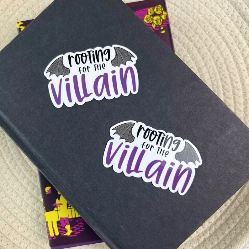 May include: Two white stickers with the text "rooting for the villain" in black and purple, with bat wing accents. The stickers are on a dark gray book. The background is a light beige woven surface.