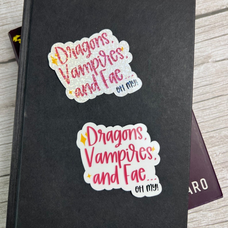May include: Two white stickers with pink and gold glitter lettering. The stickers say "Dragons, Vampires, and Fae... Oh my!"