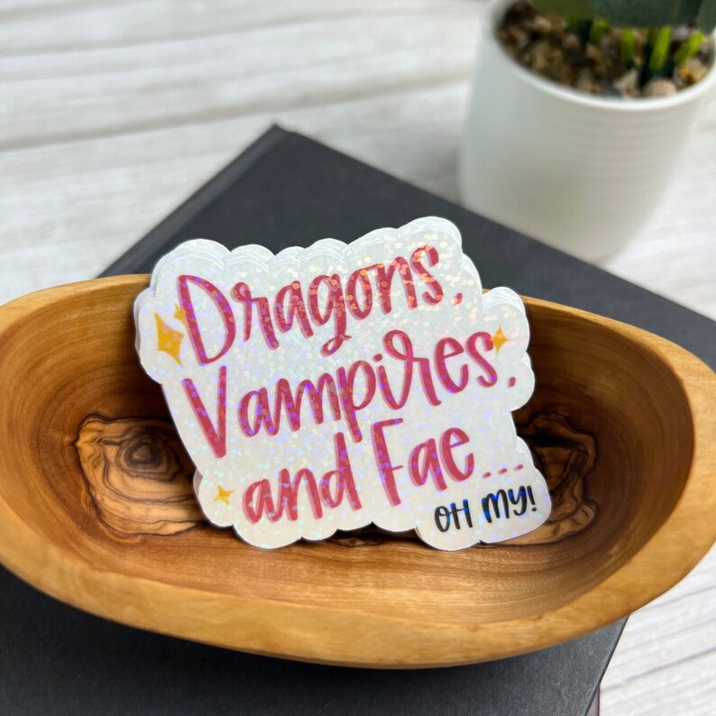 May include: A white sticker with pink glitter text that reads "Dragons, Vampires, and Fae... Oh my!" The sticker is in a wooden bowl.