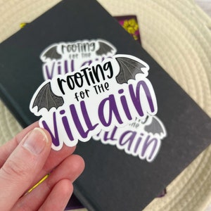 May include: A white sticker with the text "Rooting for the Villain" in black and purple, with bat wing accents. The sticker is held in front of a black notebook.