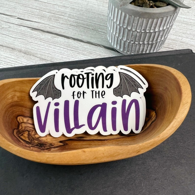 May include: A white sticker with the text "rooting for the villain" in black and purple, with bat wing accents. The sticker is in a wooden bowl.