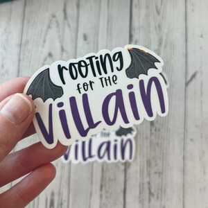 May include: A white sticker with the text "rooting for the Villain" in black and purple, with bat wing illustrations. The sticker is held by a hand.