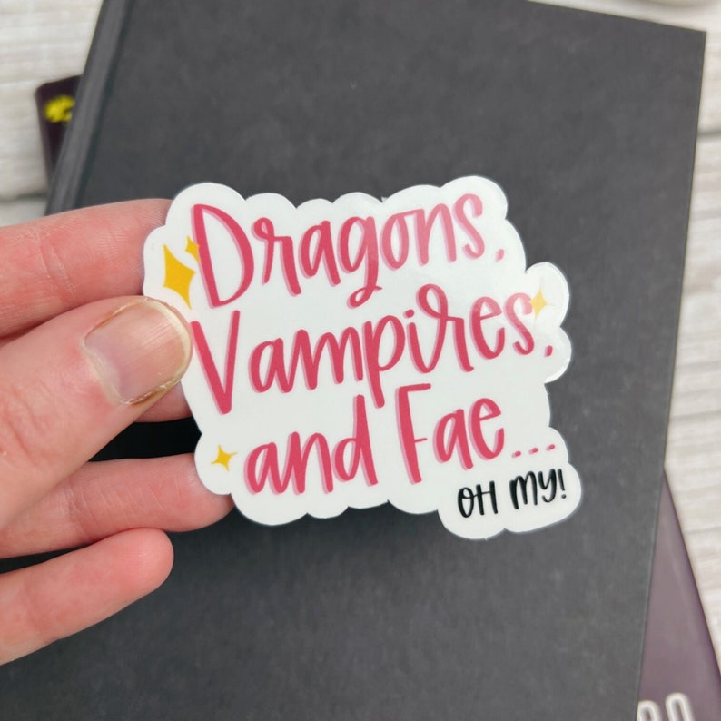 May include: White sticker with pink lettering that reads "Dragons, Vampires, and Fae... Oh my!" with yellow stars around the text.