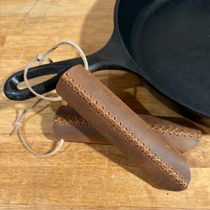 May include: A black cast iron skillet with two brown leather handle covers. The handle covers are stitched with a contrasting thread and have a loop for hanging. Kitchenware.