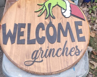 May include: A round wooden sign with the words "WELCOME grinches" in black lettering. The sign features a green hand with a red and white cuff, and a rope for hanging. The sign is placed on a gray surface.