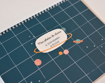 May include: A blue photo album with a white spiral binding. The cover features a white speech bubble with the text "Mes photos de classe & SOUVENIRS D'ECOLE" and a cartoon illustration of planets and stars.
