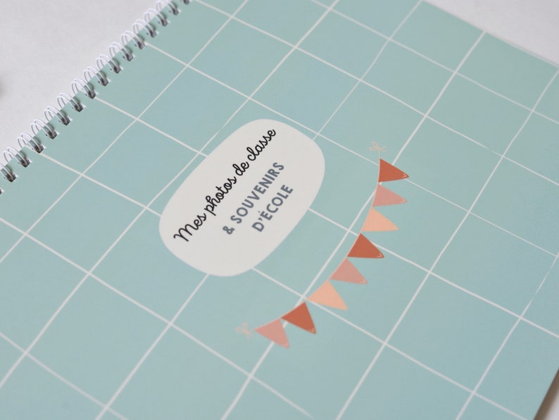 May include: A light blue photo album with a white grid pattern and a white speech bubble with the text "Mes photos de classe & souvenirs d'&eacute;cole" in black. A string of peach and brown triangle flags hangs from the top of the speech bubble.