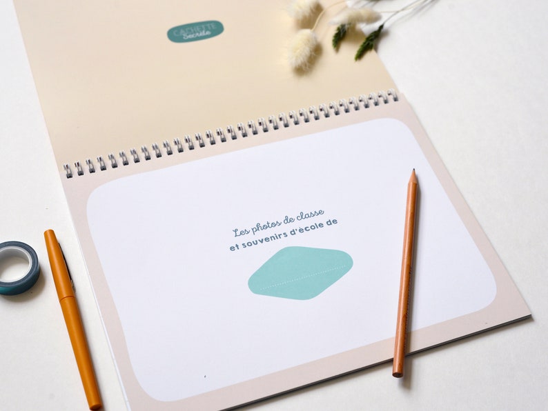 May include: A spiral bound book with a light beige cover and a teal blue graphic. The cover text reads "Les photos de classe et souvenirs d'&eacute;cole de".