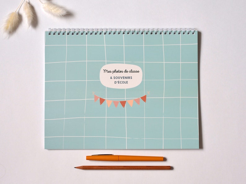 May include: A blue and white gridded notebook with a spiral binding. The notebook has the text "Mes photos de classe & SOUVENIRS D'ECOLE" and a string of colorful flags.