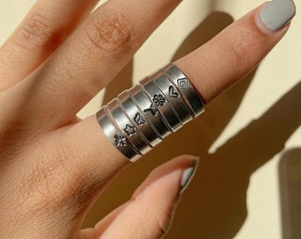 May include: A stack of silver rings with different symbols engraved on them, including a star, a flower, a clover, a butterfly, a heart, and a square. The rings are worn on a finger.