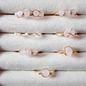 May include: A collection of handmade rings with rose quartz stones and gold-colored wire settings. The rings are displayed in a white jewelry box, showcasing various designs, including bands with multiple stones and single-stone settings.