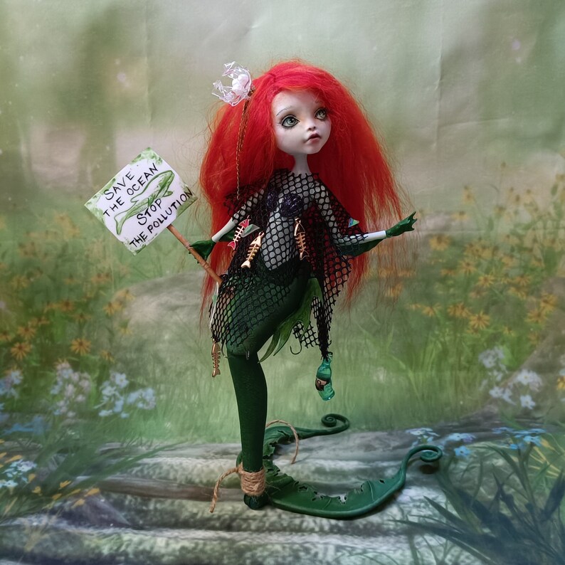 May include: A handmade doll with red hair and green skin, dressed as a mermaid. The doll holds a sign that reads "Save the Ocean Stop the Pollution." The doll is adorned with fish bones and a net. The background is a blurred image of a natural setting.