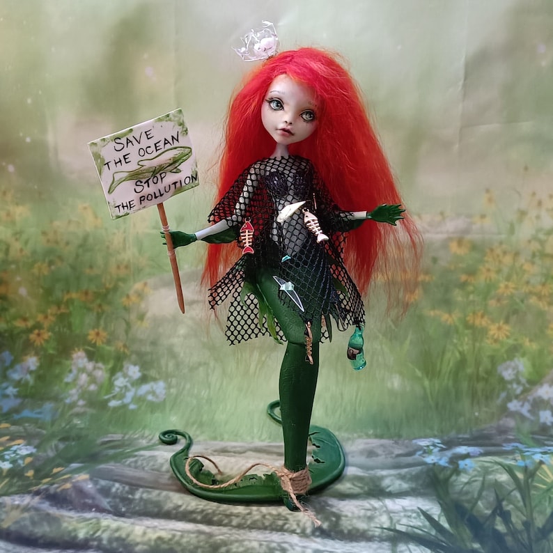 May include: A handmade doll with red hair and green skin, wearing a black net dress and holding a sign that reads "Save the Ocean Stop the Pollution." The doll is decorated with fish bones and plastic debris.