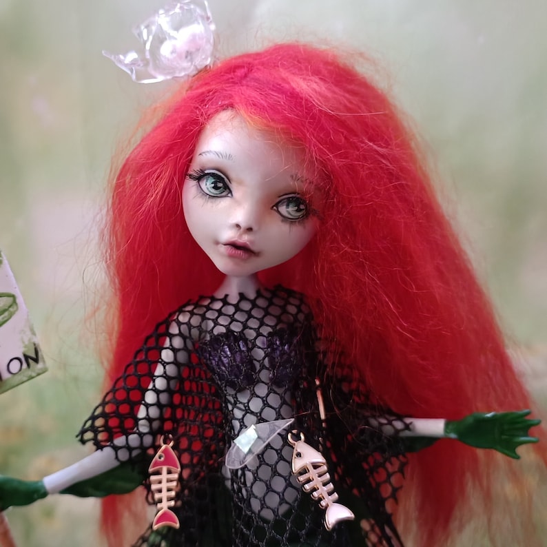 May include: A custom doll with red hair, pale skin, and green eyes. The doll wears a black mesh top, a purple bodice, and gold fishbone earrings. A clear plastic crown sits atop the doll's head. The doll has green gloves.
