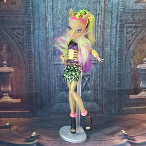 May include: A Monster High doll with a tan complexion, wearing a black and green patterned skirt, a yellow and black top, and black platform heels. The doll has green and purple hair, and is posed on a silver stand.